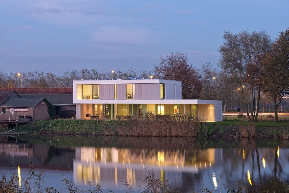 Modern huis aan het water bij schemering met grote ramen en warme verlichting die weerspiegelt in het stille meer